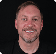 Smiling man with short brown hair and beard against black background.
